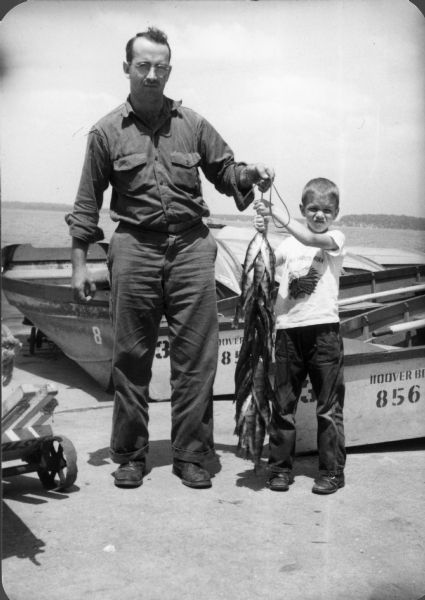 A man and boy stand on a landing in front of two boats with water and a far shoreline in the background. They are holding up their catch of fish.