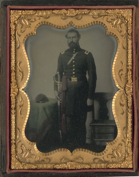 Quarter plate full-length military portrait of Harlow Milton Waller, facing forward with right hand holding his sword at his side. The portrait is hand tinted. A flat-topped cap is placed on a green cloth covered table at his right. A red sash hangs over his belt to the right of the buckle. The buttons, sword sheath, buckle and shoulder boards are tinted gold. 