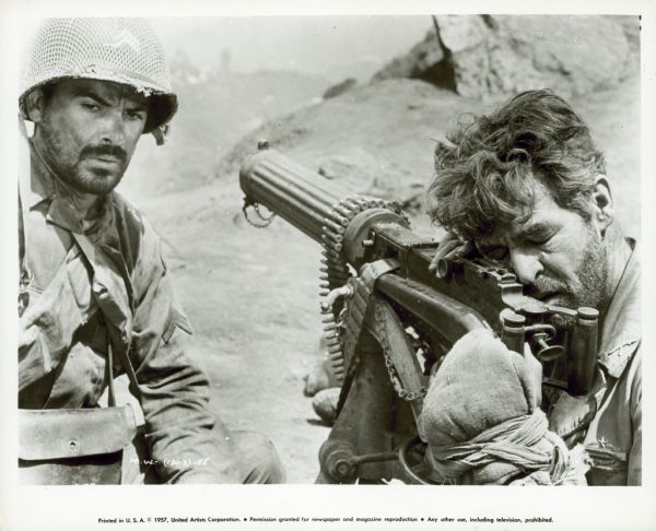 Robert Ryan and Phillip Pine are seen in a still from the film "Men in War." Exhausted and with his eyes closed, Ryan rests his head against a Vickers machine gun. Pine stares blankly.