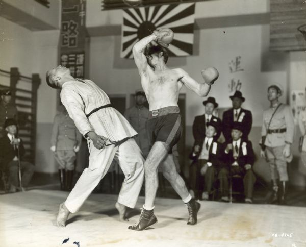 Robert Ryan, as Lefty O'Doyle, is seen boxing against a man in a white martial arts uniform. Ryan has hit the man who is falling backwards. Japanese men in suits and military uniforms can be seen sitting and standing in the background.