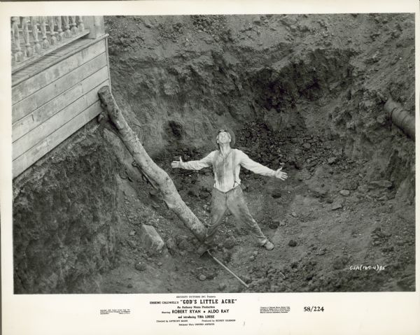 Robert Ryan, as Ty Ty Walden, stands in a large hole he has dug. He looks up to the sky with his arms stretched out. There is a large log propped up against the house next to him.