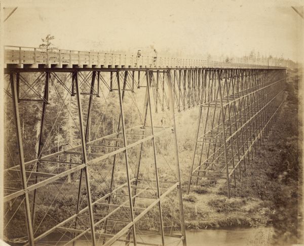 Elevated view of the Wiscsonsin Central's bridge over the White River, about six miles south of Ashland. The bridge was 1600 ft. long and 110 ft. above the water. Two people are standing on top of the bridge leaning on the railing. Another man is perched below them on a support beam. The perspective of this photograph illustrates well why the White River Bridge was considered a marvel of railroad engineering. The bridge was dismantled and sold for scrap in 1909.