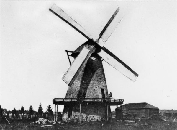 Wind powered stone grist mill. Two men stand on a wood balcony near the open doorway of the windmill.