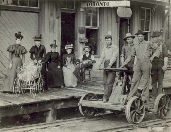 Employes of the Chicago, Milwaukee & St. Paul Railway and their wives, Mr. and Mrs. N.J. Edwards, W.H. Lenshan, E.B. McLaird, Alf. Hetig, and George McLaird, Sr. Four men are posing on a railroad handcar on the railroad tracks in front of the Toronto railroad station. Four women, an infant in a baby carriage, and one man, pose on the railroad platform behind them.