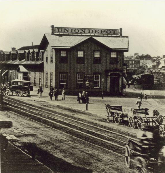 Slightly elevated view over railroad tracks of the Union Depot, erected in 1866 on Reed Street. Horse-drawn wagons and cabs are parked nearby,  and pedestrians are on the platform. A railroad car can be seen on another set of railroad tracks curving towards buildings on the right. Caption reads: "Milwaukee & St. Paul Railroad Company built Brookfield to Milwaukee in 1864 and used a small frame structure for a depot until erection of the Reed Street 'Union Depot' in 1866. The temporary building was moved to the west side of the track and used for an emigrant depot, — the new building was placed on the site occupied by the building that was moved."