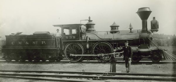 View across railroad tracks of two men posing near the right side of locomotive no. 539 on another set of railroad tracks. The locomotive was built by the Schenectady Locomotive Works for the Chicago, Milwaukee, and St. Paul Railway.
