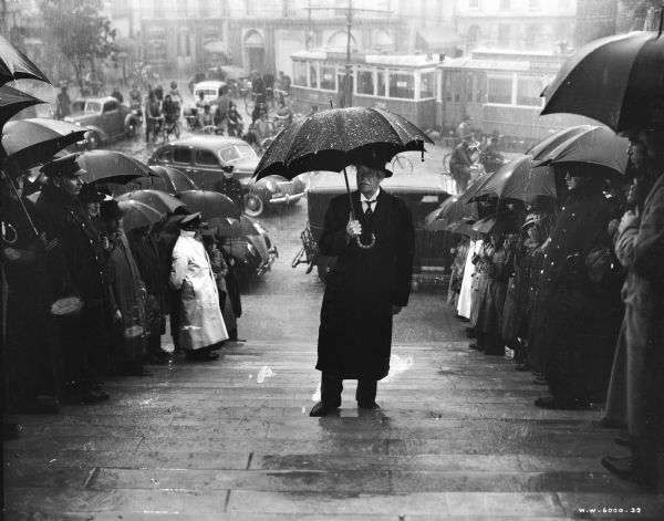 Albert Basserman is seen on the set of the film "Foreign Correspondent." He is walking up a flight of stairs in the rain holding an umbrella. A crowd of people holding umbrellas are lined up along both sides of the steps.