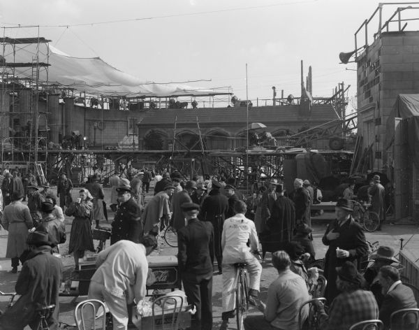 Outdoor view of the Amsterdam Square set of the film "Foreign Correspondent." Scaffolding and lights are in the background, and many members of the cast and crew are sitting and standing around the set.