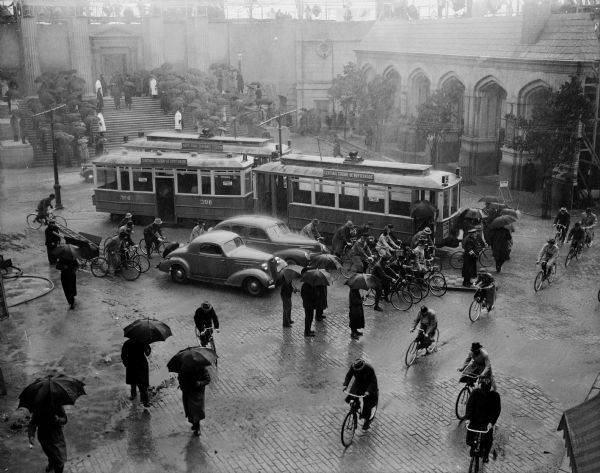 Elevated scene still for the film "Foreign Correspondent." Pedestrians are walking with umbrellas down the sidewalk in the rain. On the street people are riding bicycles near two automobiles and two streetcars. In the background a crowd of people stand on the steps in front of a building.