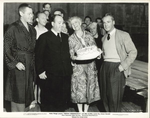 Alfred Hitchcock stands next to Gertrude Hoffman who is smiling and holding a birthday cake. Walter Wanger stands to the right of the woman, and Herbert Marshall, wearing a robe and holding a cigarette, stands to the left of Hitchcock. A group of people stands behind them.  The photo was taken on the set of the film "Foreign Correspondent".
