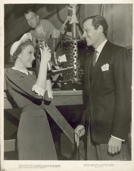 Laraine Day takes a photograph of Joel McCrea; both are smiling. Both are dressed very nicely. In the background a crew member looks at them from a raised platform.