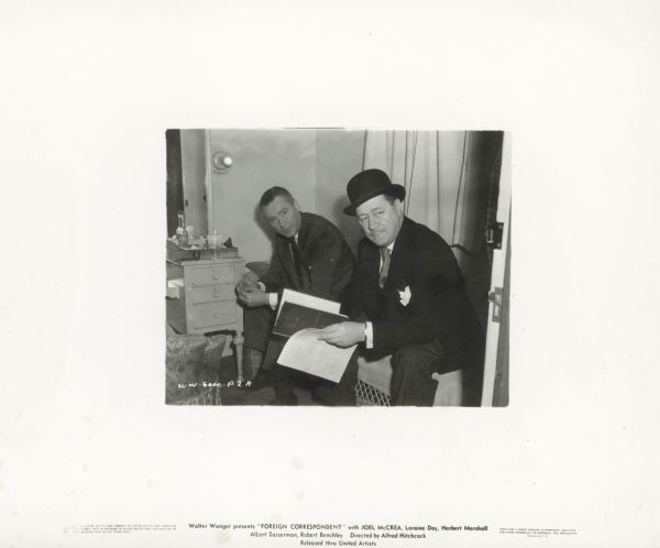 Herbert Marshall and Robert Benchley sit next to each other on the set of the film "Foreign Correspondent." Both wear suits; Benchley holds a script and is wearing a bowler.