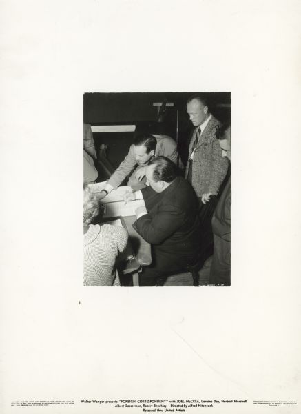 Slightly elevated view of Alfred Hitchcock sitting and sketching a storyboard on the set of the film "Foreign Correspondent." Three men and a woman stand around him.