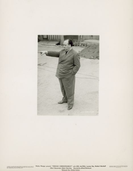 Alfred Hitchcock stands on the set of the film "Foreign Correspondent." He has his left hand in his pants pocket and is pointing at something with his right hand.