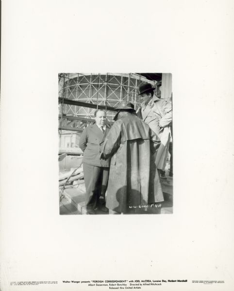 Alfred Hitchcock stands with Joel McCrea and another man on the set of the film "Foreign Correspondent." McCrea wears a suit, trench coat and bowler; the other man wears a rain coat and hat and has his back to the camera.