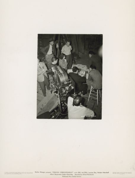 An overhead photograph from behind of Alfred Hitchcock sitting and drawing a storyboard on the set of the film "Foreign Correspondent." He is surrounded by seven men and one woman. A motion picture camera is positioned behind Hitchcock.