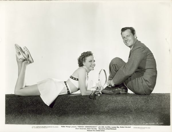 Publicity photo of Joel McCrea and Laraine Day for the film "Foreign Correspondent." McCrea wears a suit and sits with his arms around his legs. Day wears a bikini top, skirt, and sandals. She is lying on her stomach with her feet in the air and holds a tennis racket.