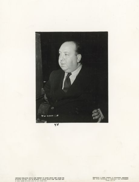 Alfred Hitchcock stands with his hands on his hips. He is wearing a suit and looking off to the side.