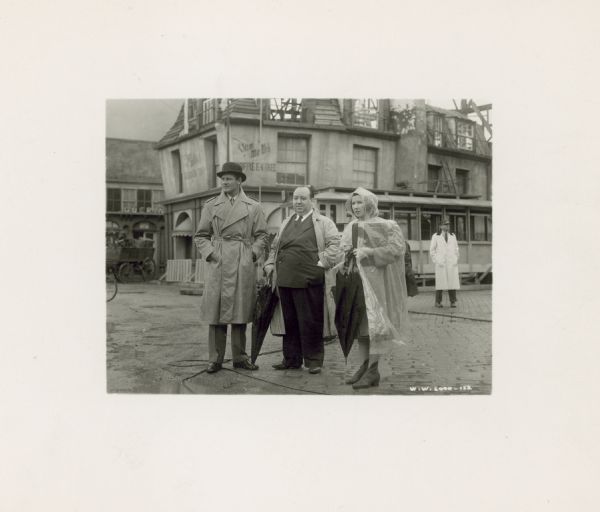 Alfred Hitchcock and Joel McCrea stand on the set of the film "Foreign Correspondent" with an unidentified woman wearing a rain poncho. Hitchcock and the woman hold umbrellas. McCrea wears a trench coat and bowler hat.