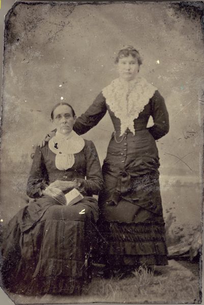 Studio portrait in front of a painted backdrop of two women. The woman on the left wears eyeglasses and a white collar and is sitting in a chair with a book in her lap. The woman on the right is wearing a lace collar over her dress and is standing. She has her hand resting on the sitting woman's shoulder.