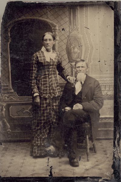 Studio portrait in front of a painted backdrop of a man and a woman. The woman is standing on the left and has her hand resting on the shoulder of the man on the right, who is sitting.