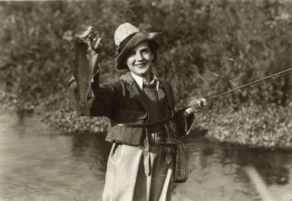 A woman wearing waders and a hat holds up a trout in her right hand and a fly-fishing rod in her left hand. Attached to her hat are fly-fishing flies, and she is wearing a wicker creel around her waist.
