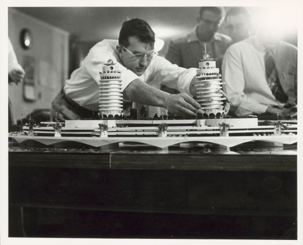 An architect sets a tower on an artitectural model of the Monona Terrace. Other men watch in the background.