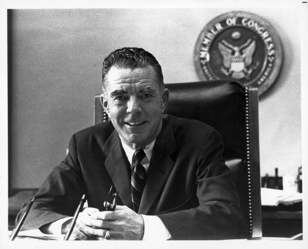 A casual portrait of John W. Byrnes seated at a desk, with a "Member Of Congress" plaque on the wall behind him.