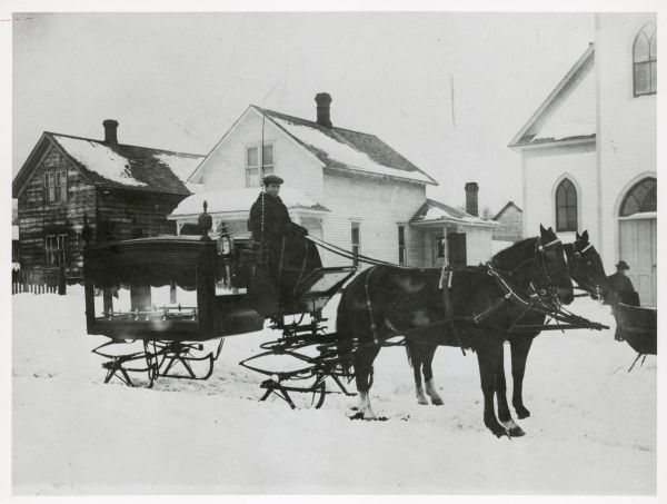 Martin Keefe, owner of the livery stable, drives a horse-drawn hearse, which has sled runners. The hearse was for the funeral of Mrs. Tom Hill. The buildings in the background are the homes of Nick Rodesch and Peter Napierala, and the Presbyterian Church.