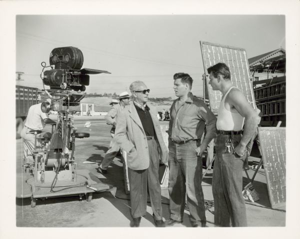 Walter Wanger talks with actors Neville Brand and Leo Gordon outdoors on the set of the film "Riot in Cell Block 11." A large movie camera on wheels is on the left.