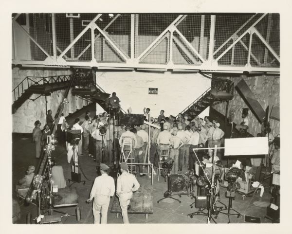 Elevated view of the set for the film "Riot on Cell Block 11." There is a large group of men in the center back, and movie cameras, lighting equipment, and more men in the foreground. There are flights of stairs on the right and left. A catwalk runs across the set above the actors. On the back wall a sign reads: "Notice: Prisoners must not go on balconies without permission."