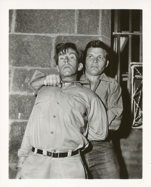 Actor Neville Brand stands behind actor Whit Bissell while holding a knife to his throat. They are on the set of the film "Riot in Cell Block 11."