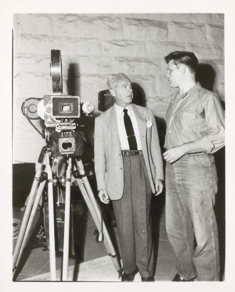 Walter Wanger stands on the left while talking with actor Neville Brand, on the right. On the far left is a movie camera. They are on the set of the film "Riot on Cell Block 11."