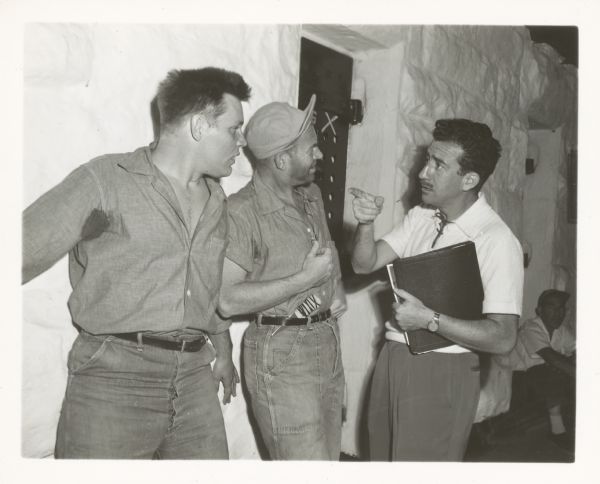 Actor Neville Brand, on the left, and another actor in the center with a knife in his belt, talk with director Don Siegel, far right. They are on the set of the film "Riot in Cell Block 11." Siegel holds a binder and points at Brand. The two actors are standing against a wall.