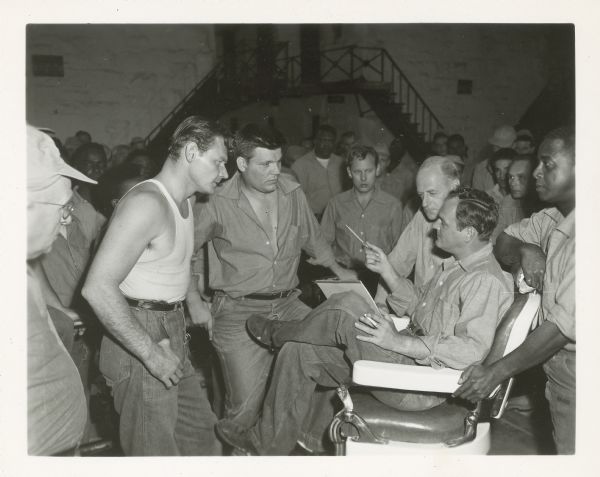 Leo Gordon, Neville Brand and Joel Fluellen stand in front of Robert Osterloh, sitting, who is holding a pencil in one hand and a cigarette in the other. A large group of men are standing in the background on the stage set with stairways.