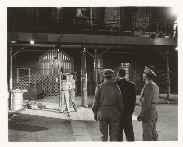 Long shot of a scene from the film "Riot in Cell Block 11." In the background, Neville Brand holds a knife to the throat of Whit Bissell. Two guards and a warden stand in the foreground with their backs to the camera.
