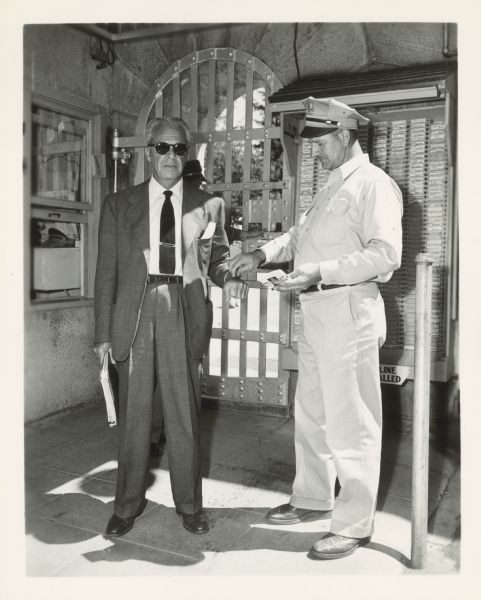 Walter Wanger gets his hand stamped by a prison guard. He is wearing a suit and sunglasses and holds a binder.