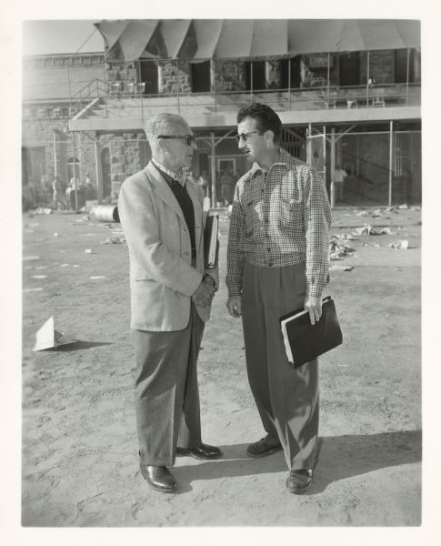 Walter Wanger and Don Siegel talk on the set of the film "Riot in Cell Block 11." They are standing outside in the prison yard. They both hold binders.
