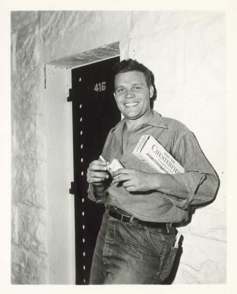 Neville Brand leans against a wall outside of a prison cell and smiles. He is pulling out a cigarette from a cigarette pack he holds in his hands. There is a carton of Chesterfield cigarettes under his arm.