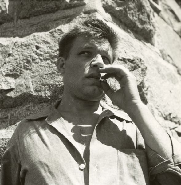 Contact print of actor Neville Brand smoking a cigarette. He is outside on the set of the film "Riot in Cell Block 11" in front of the corner of a stone wall.