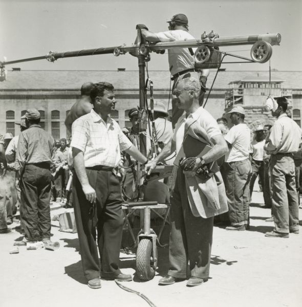Walter Wanger is seen talking to a man on the set of the film "Riot in Cell Block 11." A large microphone boom on wheels is directly behind them. They are in the yard of a prison.