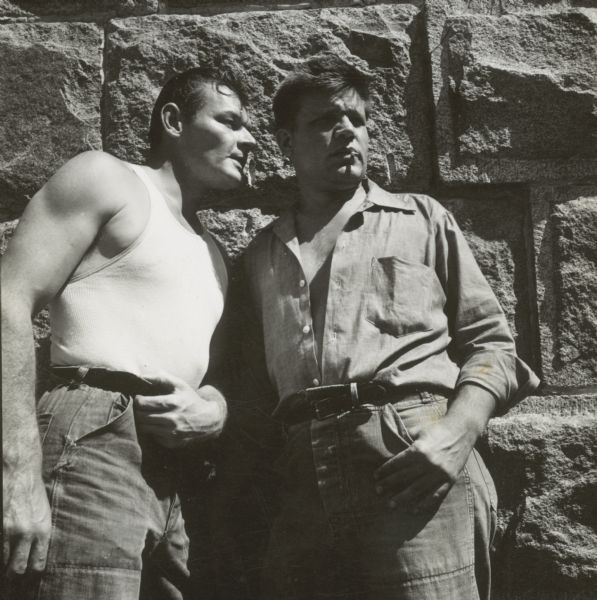 Neville Brand and Leo Gordon are seen outside leaning against a stone wall on the set of the film "Riot in Cell Block 11." Gordon is whispering into Brand's ear.