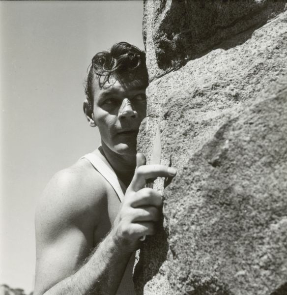 Leo Gordon holds a knife and looks around a stone wall. He is outside on the set of the film "Riot in Cell Block 11."