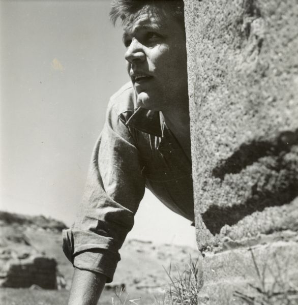 Neville Brand kneels on the ground and looks out from behind a stone wall on the set of the film "Riot in Cell Block 11."