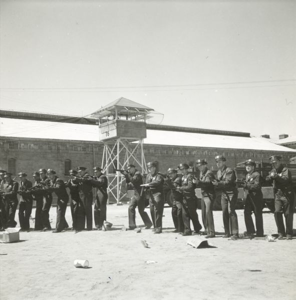 A line of police officers pointing rifles are seen on the set of the film "Riot in Cell Block 11." They are outside in the yard of a prison. A guard tower can be seen in the background.