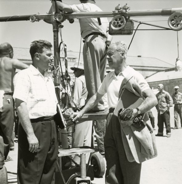 Walter Wanger is seen talking to a man on the set of the film "Riot in Cell Block 11." They are standing outside in the yard of a prison. Wanger has his hand on a microphone boom and holds his coat and binder in the other arm.
