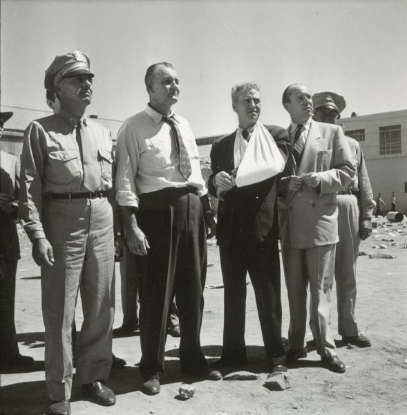 Emile Meyer and Frank Faylen and three other men are seen on the set of the film "Riot in Cell Block 11." They are standing outside in the yard of a prison. Faylen has his arm in a sling.
