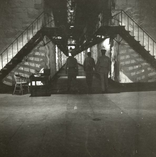 Long shot of two wardens escorting a prisoner back to a cell on the set of the film "Riot in Cell Block 11." They are walking underneath two metal staircases.
