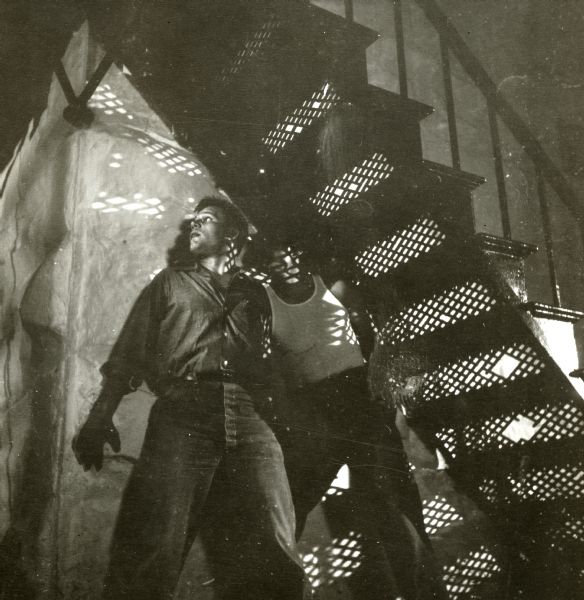 Neville Brand and Leo Gordon are seen hiding underneath some metal stairs on the set of the film "Riot in Cell Block 11."