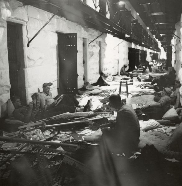 Prisoners are seen sitting outside of cells on the set of the film "Riot in Cell Block 11." There is debris on the floor. A prisoner wearing a hat and glasses can be seen leaning up against a wall and mattress reading.